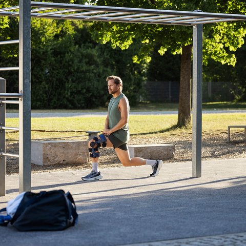 Ein Mann mit Knieorthese führt draussen einen Ausfallschritt unter einer Kletterstruktur aus. Im Vordergrund liegt eine Sporttasche, im Hintergrund sind Bäume und ein Parcours sichtbar.