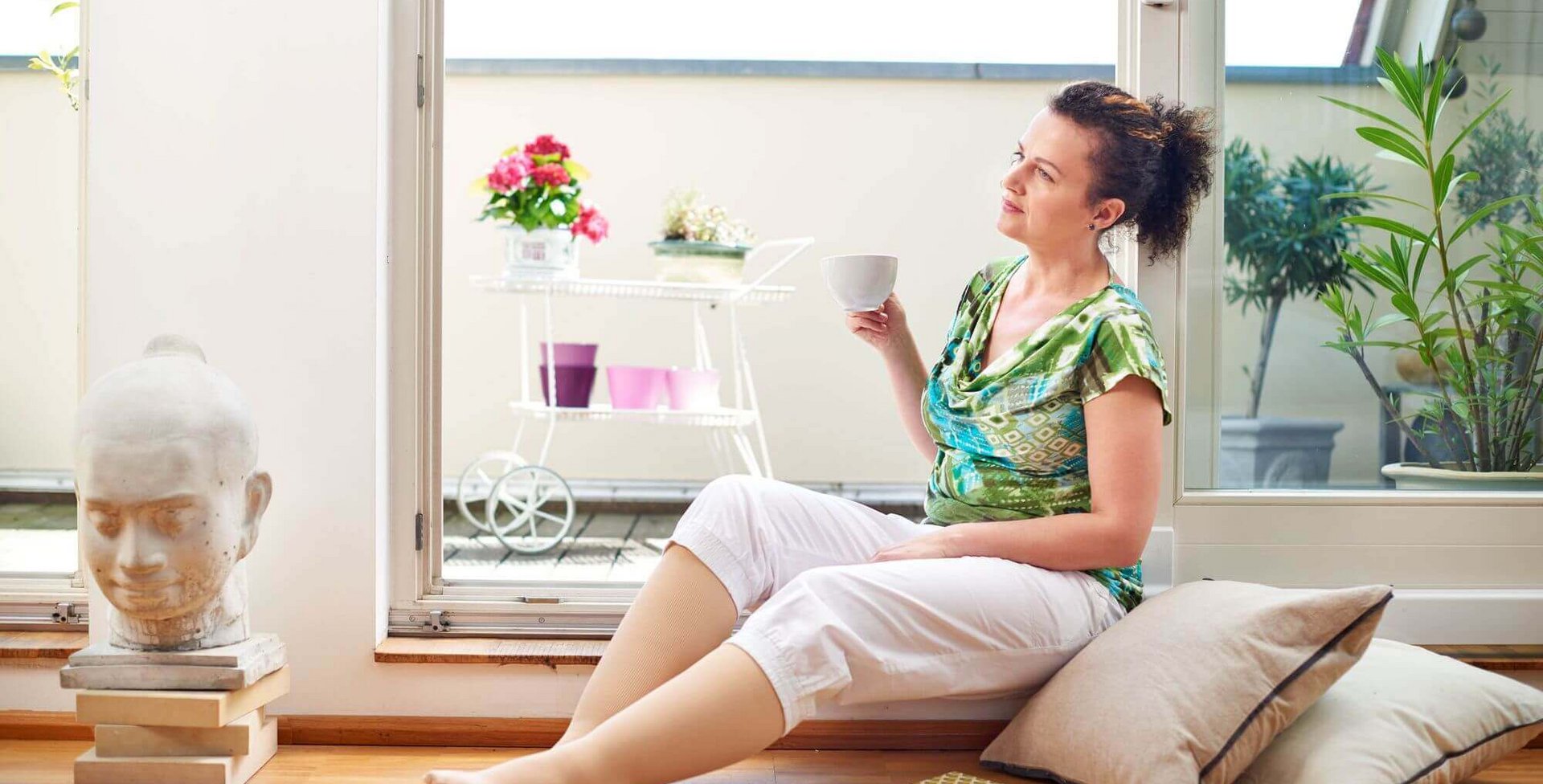 Eine Frau sitzt, an Kissen angelehnt, auf dem Fußboden in einem Wohnzimmer und trinkt genüsslich eine Tasse Kaffee. Sie trägt eine VenoTrain curaflow Beinversorgung von Bauerfeind. Dies ist ein Kompressionsstrumpf zur gezielten Ödemtherapie am Bein.