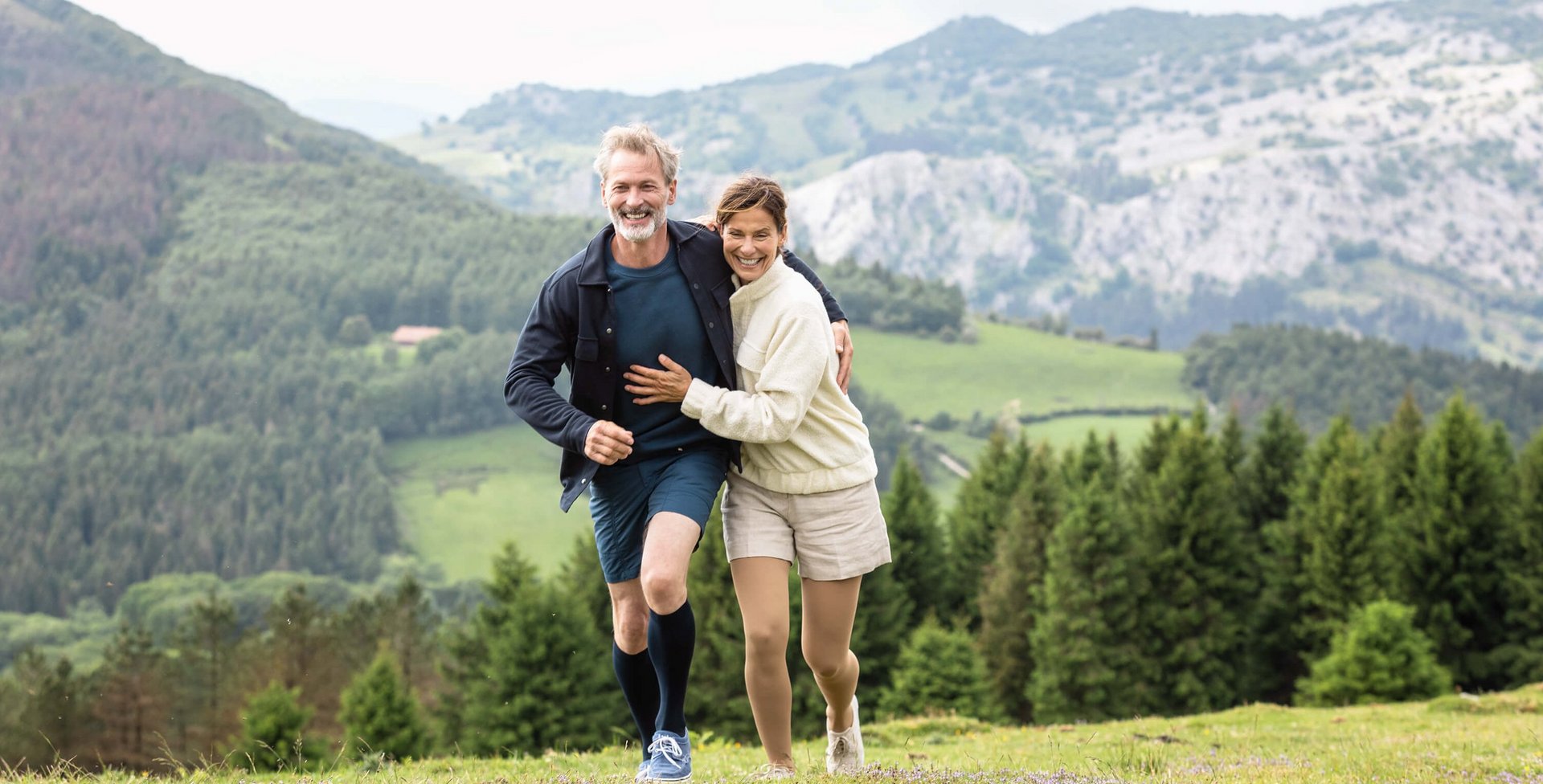 Ein Mann und eine Frau laufen Arm in Arm in einer Berglandschaft über eine Wiese. Beide tragen VenoTrain impuls Kompressionsstrümpfe von Bauerfeind. Diese helfen bei Besenreisern.