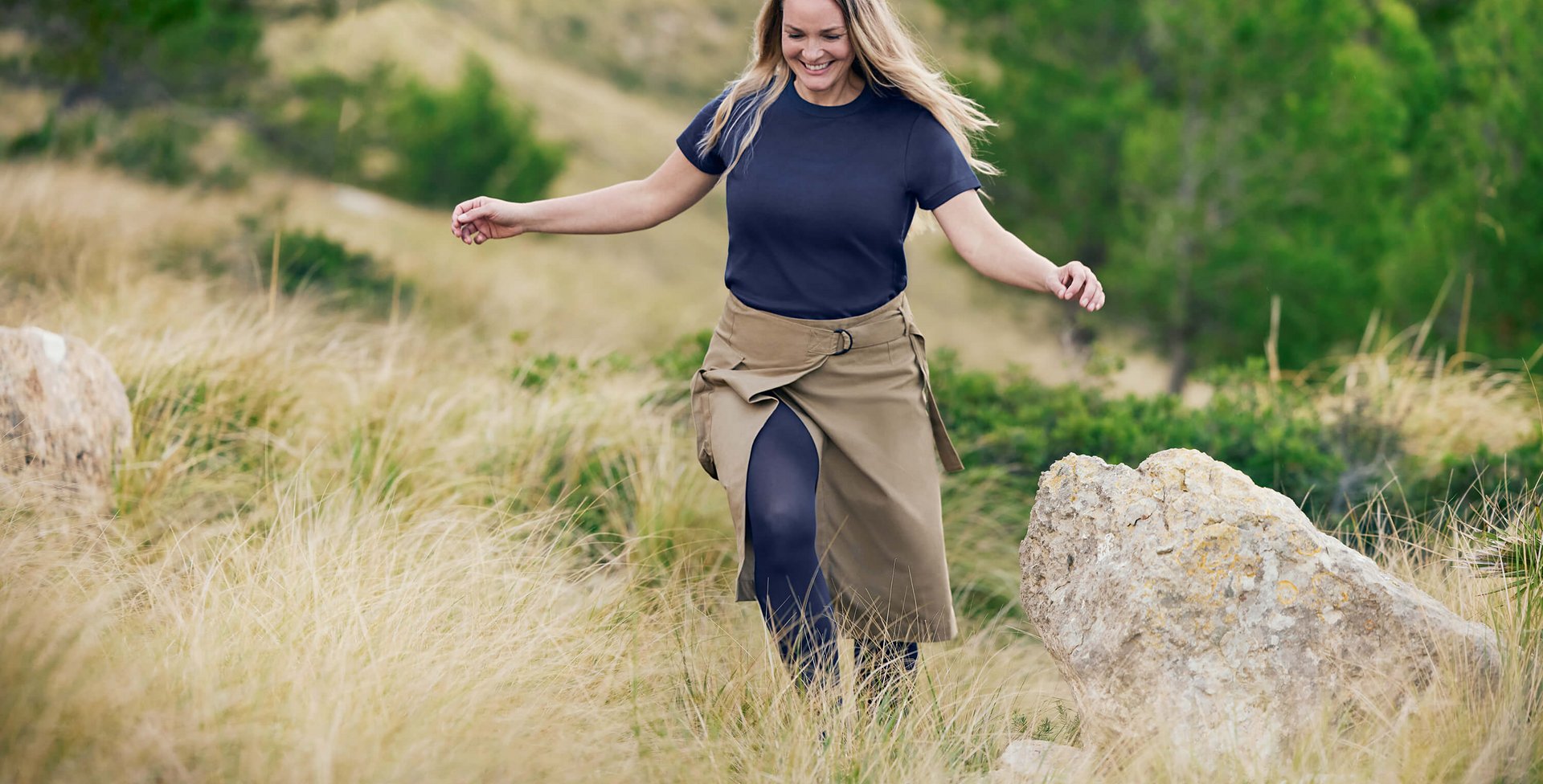 Eine lächelnde Frau mit langen, offenen Haaren geht leichtfüßig durch eine hügelige Naturlandschaft mit hohem Gras und Felsen. Sie trägt ein lässiges dunkelblaues T-Shirt, einen khakifarbenen Wickelrock und VenoTrain Kompressionsstrümpfe in Dunkelblau.