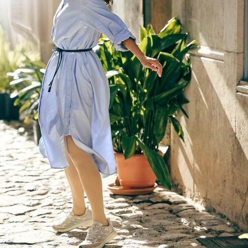 Eine lachende Frau mit wehenden Haaren dreht sich spielerisch in einer sonnigen Altstadtgasse. Sie trägt ein locker geschnittenes hellblaues Hemdblusenkleid, eine beigefarbene Cap und VenoTrain Kompressionsstrümpfe in einem natürlichen Ton, kombiniert mit sportlichen Sneakern.