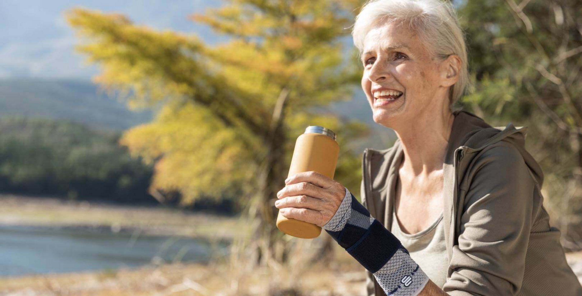 Auf dem Bild ist eine ältere Frau mit Getränk an einem See zu sehen, die beim Wandern eine ManuTrain Handgelenkbandage trägt, um Schmerzen im Handgelenk zu lindern.