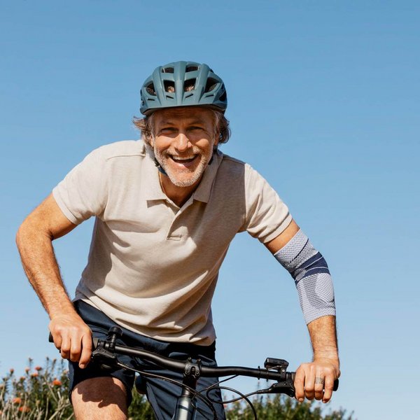 Mann mit Fahrradhelm fährt bei klarem Himmel lächelnd Fahrrad, trägt die EpiTrain Ellenbogenbandage von Bauerfeind am linken Arm zur Unterstützung des Gelenks