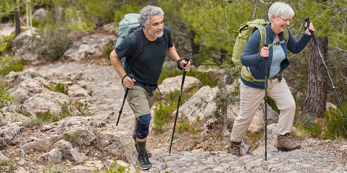 Ein Mann und eine Frau wandern auf einem steinigen Weg durch einen Wald. Beide tragen Wanderausrüstung mit Rucksäcken und Wanderstöcken. Der Mann trägt eine GenuTrain OA Knieorthese am rechten Knie, die seine Bewegung beim Anstieg unterstützt. 