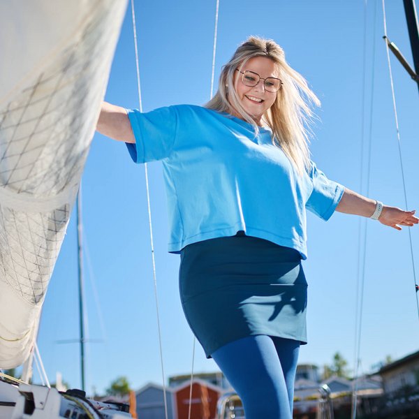 Eine Frau mit blondem Haar, die ein blaues T-Shirt und eine blaue Kompressionsstrumpfhose trägt, steht lächelnd auf einem Boot und balanciert ihre Arme aus. Im Hintergrund sind Masten und Boote zu sehen, der Himmel ist klar und blau. 
