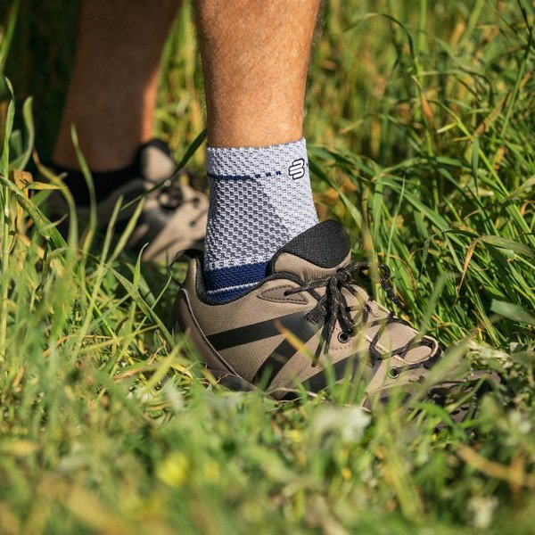 Nahaufnahme eines Fußes in einem Wanderschuh, der eine MalleoTrain Sprunggelenkbandage von Bauerfeind trägt, stehend im hohen Gras mit einem Wanderstock im Hintergrund.