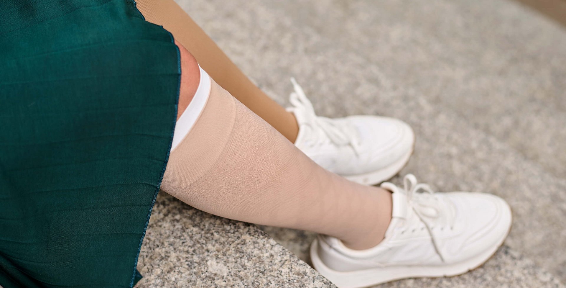 Nahaufnahme eines Beines in einem beige-farbenen Kompressionsstrumpf aus der VenoTrain Special-Linie, kombiniert mit einem grünen Kleid und weißen Sneakers. Das Bein ruht auf einer steinernen Treppenstufe.