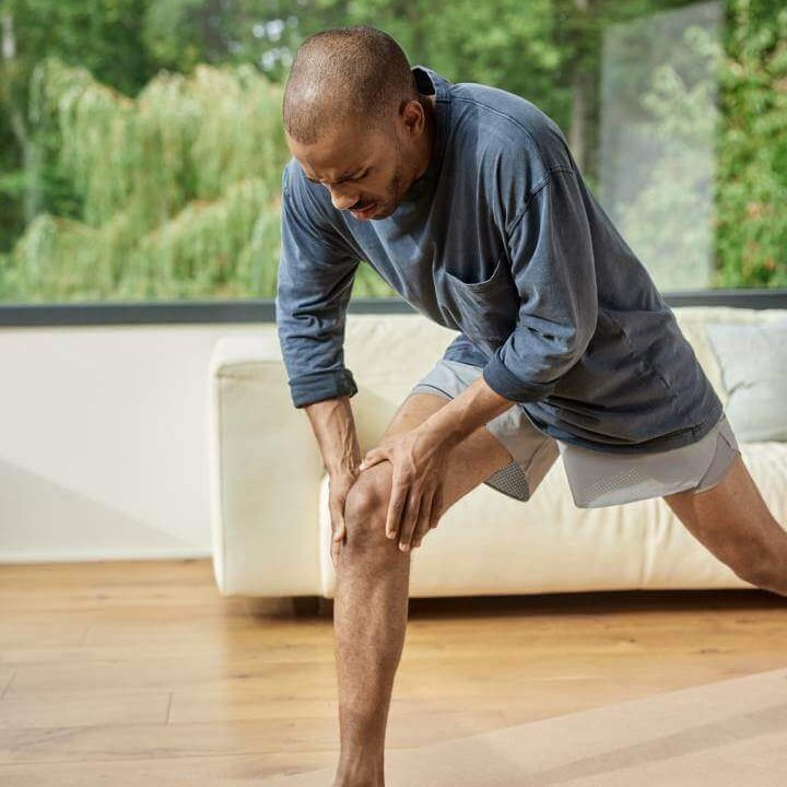 Een man grijpt tijdens de training naar zijn pijnlijke knie. Hij draagt sportieve kleding en bevindt zich in een moderne ruimte met grote ramen. Op de voorgrond zijn een waterfles, een springtouw en een yogamat te zien.