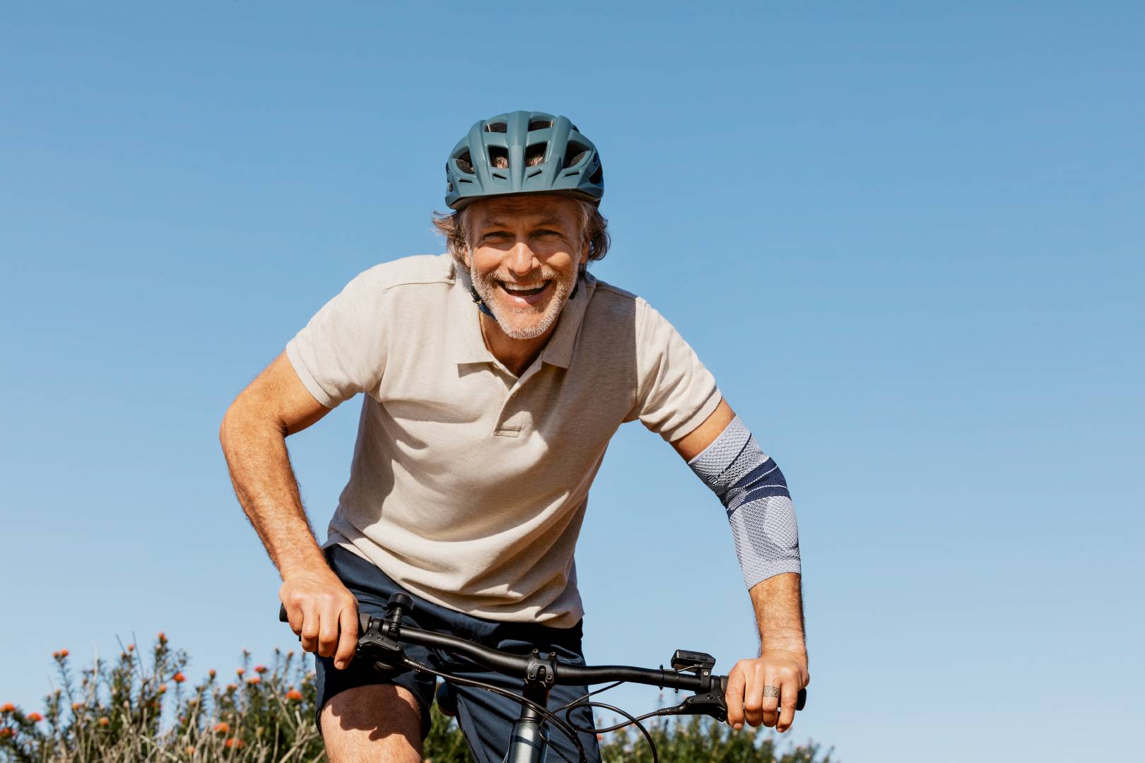 Man met fietshelm fietst bij heldere hemel lachend op zijn fiets, draagt de EpiTrain elleboogbandage van Bauerfeind aan zijn rechterarm ter ondersteuning van het gewricht.