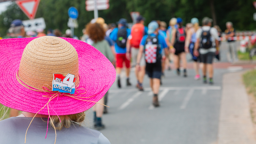 Een vrouw met een roze strohoed loopt langs een weg tijdens de Vierdaagse. Andere wandelaars zijn voor haar zichtbaar, maar slechts vaag.