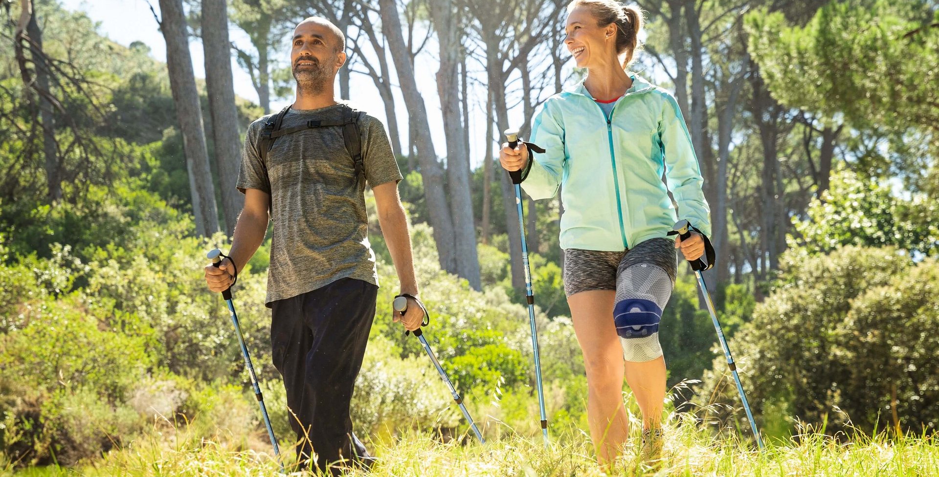 Een man en een vrouw wandelen met nordic-walkingstokken door een zonnig, groen bos. Beiden dragen sportieve kleding en de vrouw draagt een kniebandage. Ze lachen naar elkaar en ogen ontspannen.