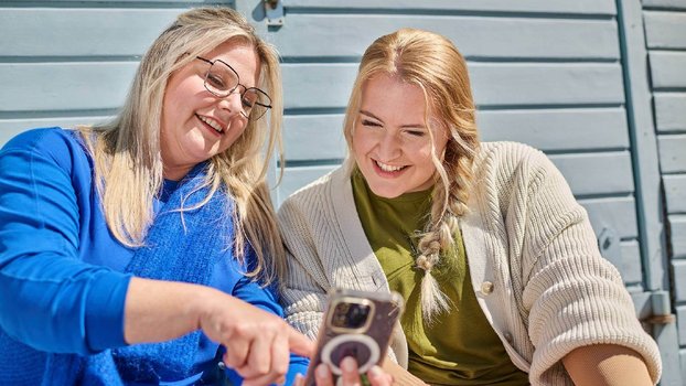 Twee vrouwen zitten buiten voor een blauwe houten wand en lachen, terwijl ze samen naar een smartphone kijken.