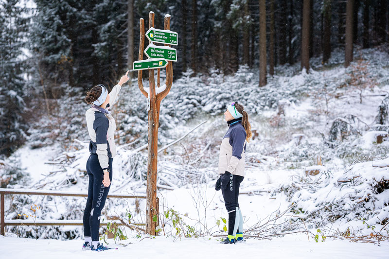 Twee vrouwen in winterhardloopkleding staan voor een houten wegwijzer in het besneeuwde bos. Een van hen wijst naar de bordjes terwijl de ander aandachtig kijkt. De wegwijzer geeft verschillende wandelroutes aan.