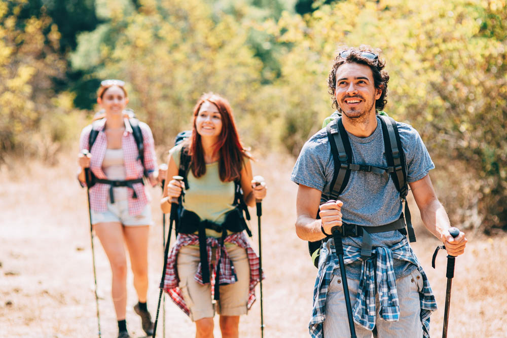 Drie wandelaars, twee vrouwen en een man, lopen vrolijk met trekkingstokken over een zonnig bospad. Ze dragen sportieve kleding en rugzakken – de nadruk ligt op natuurlijke beweging en samen buiten zijn.
