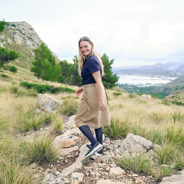Vrouw wandelt lachend door een rotsachtig landschap met uitzicht op een kust, draagt donkerblauwe compressiekousen van Bauerfeind voor de aderbehandeling, gecombineerd met een beige rok en sneakers.