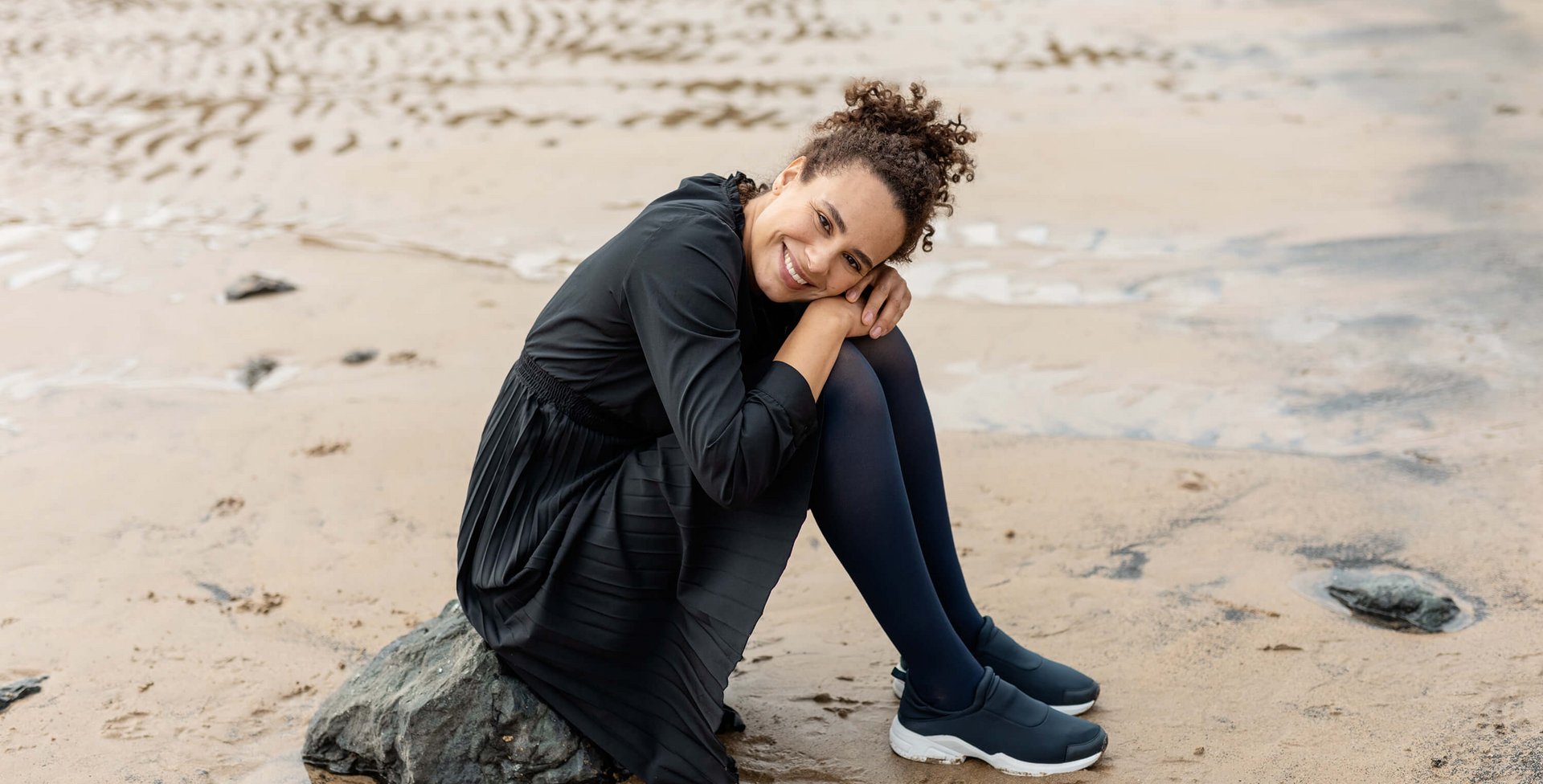 Het beeld toont een vrouw die op een kleine rots op het strand zit. Ze draagt VenoTrain soft compressiekousen van Bauerfeind.