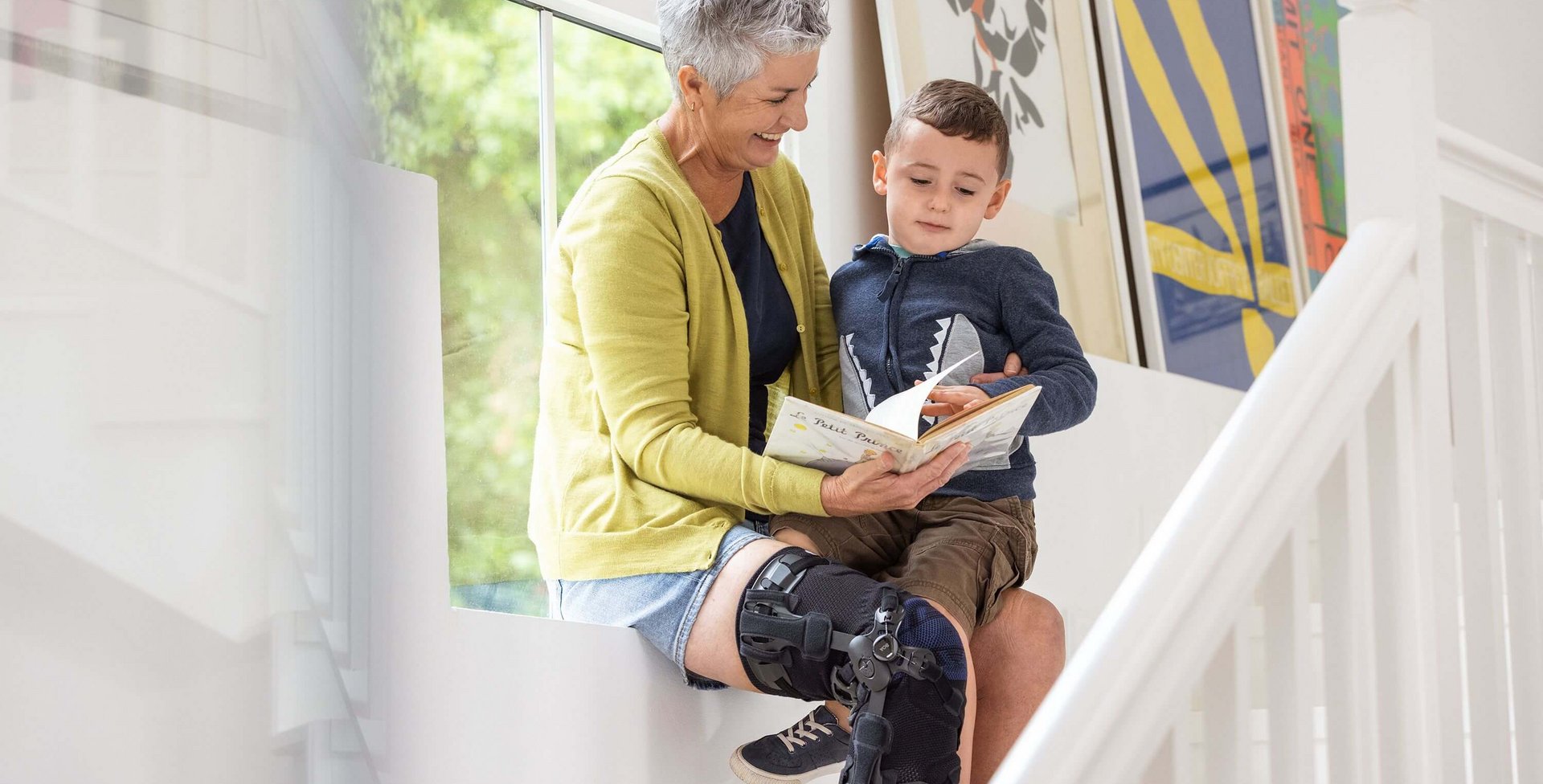 Een oudere vrouw zit op een vensterbank en leest een klein jongetje een boek voor. Ze draagt een GenuTrain OA knieorthese aan haar rechterbeen. Op de achtergrond hangen kleurrijke ingelijste foto's aan de muur.
