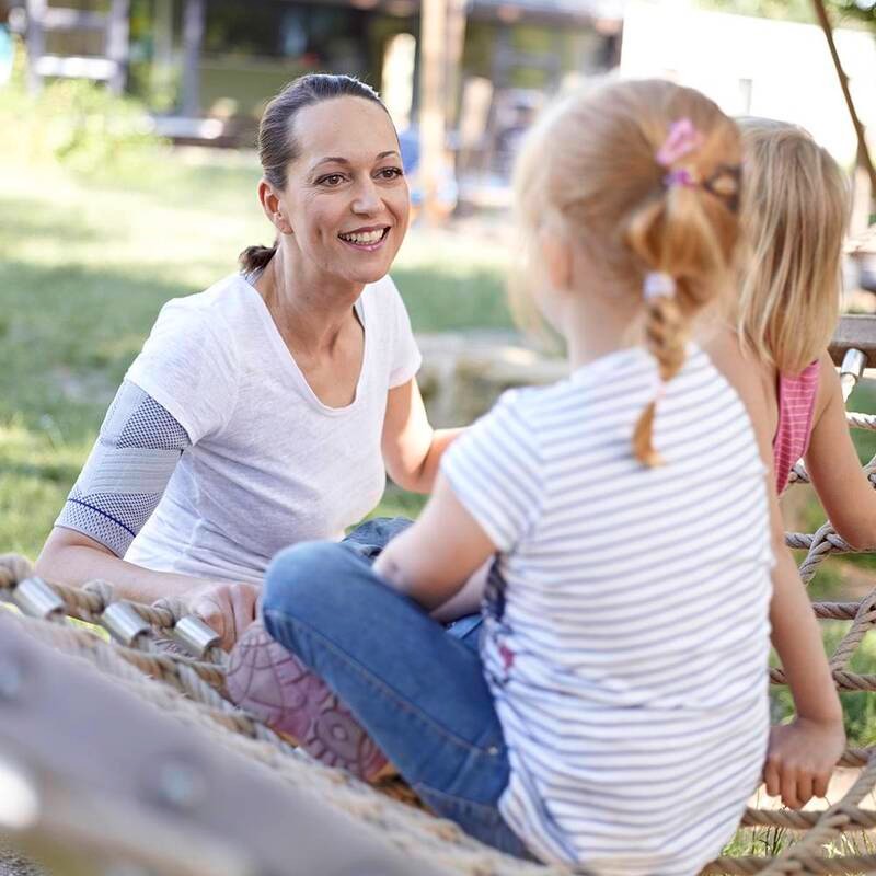 Vrouw draagt Bauerfeind OmoTrain schouderbrace tijdens spelen in het park met kinderen.