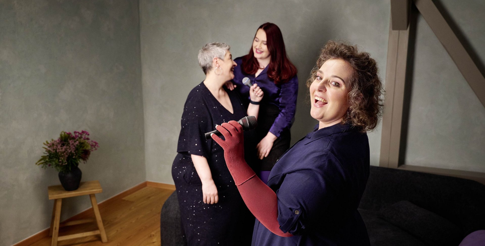 Drie vrouwen zingen samen met microfoons in een woonkamer met lichtgroene muur. De vrouw op de voorgrond draagt een donkerblauwe jurk en rode compressiehandschoenen; op de achtergrond lachen twee andere vrouwen.