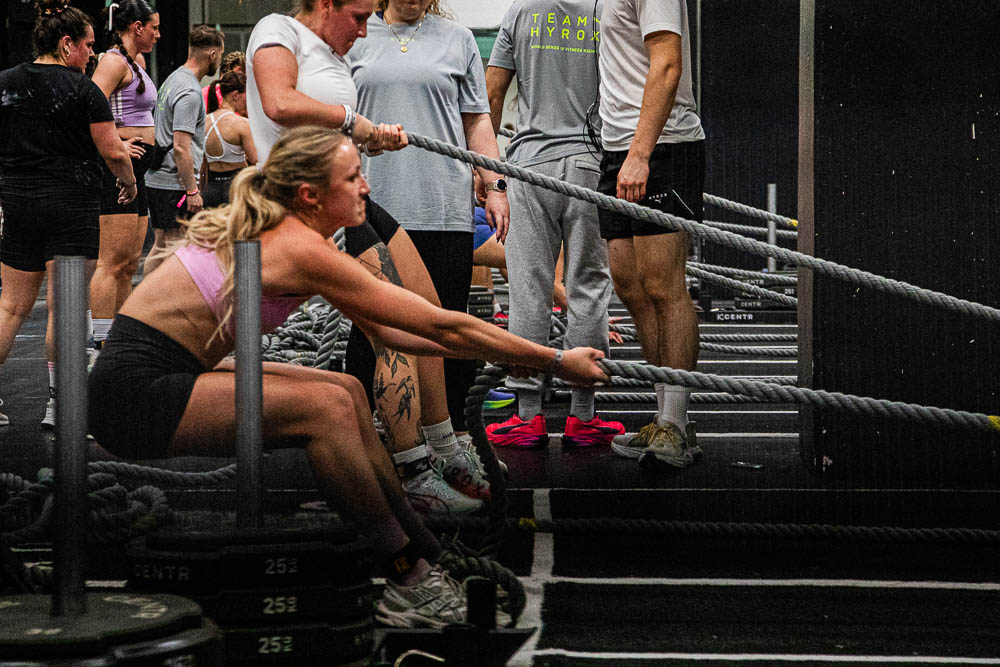 Atlete voert een sled pull uit tijdens een HYROX-wedstrijd en trekt met een lang touw een zware slee over de indoorvloer, terwijl begeleiders en andere deelnemers bij de stations op de achtergrond staan; de scène toont intensief krachtwerk,