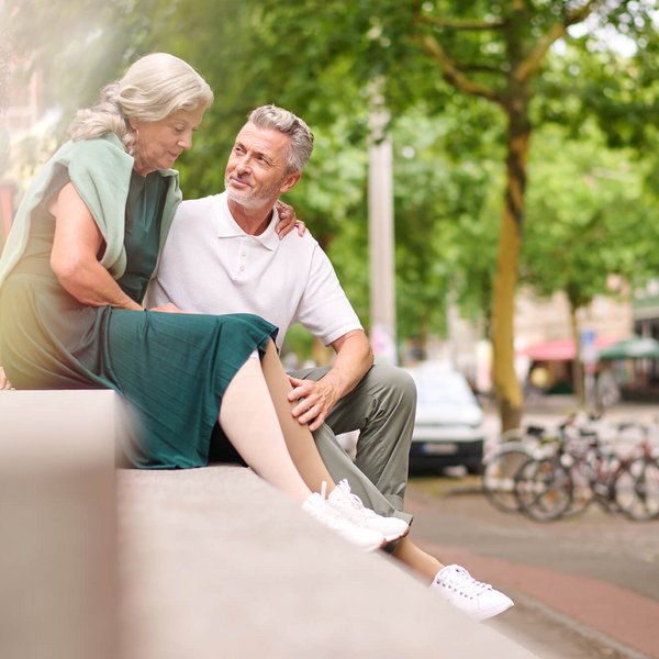 Een ouder stel zit samen op een stenen muurtje in een stedelijke omgeving, de vrouw draagt VenoTrain compressiekousen van Bauerfeind ter ondersteuning bij de wondbehandeling, gecombineerd met een groene jurk en witte sneakers.