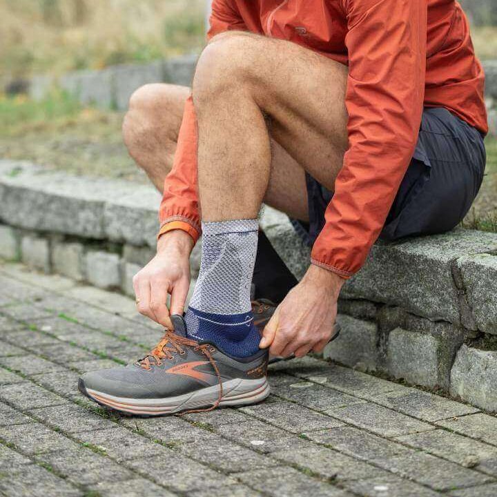 De foto toont een jonge man in de tuin. Een hond loopt om zijn benen. De man draagt een AchilloTrain enkelbandage van Bauerfeind aan zijn rechtervoet. Deze bandage ontlast gericht de achillespees.