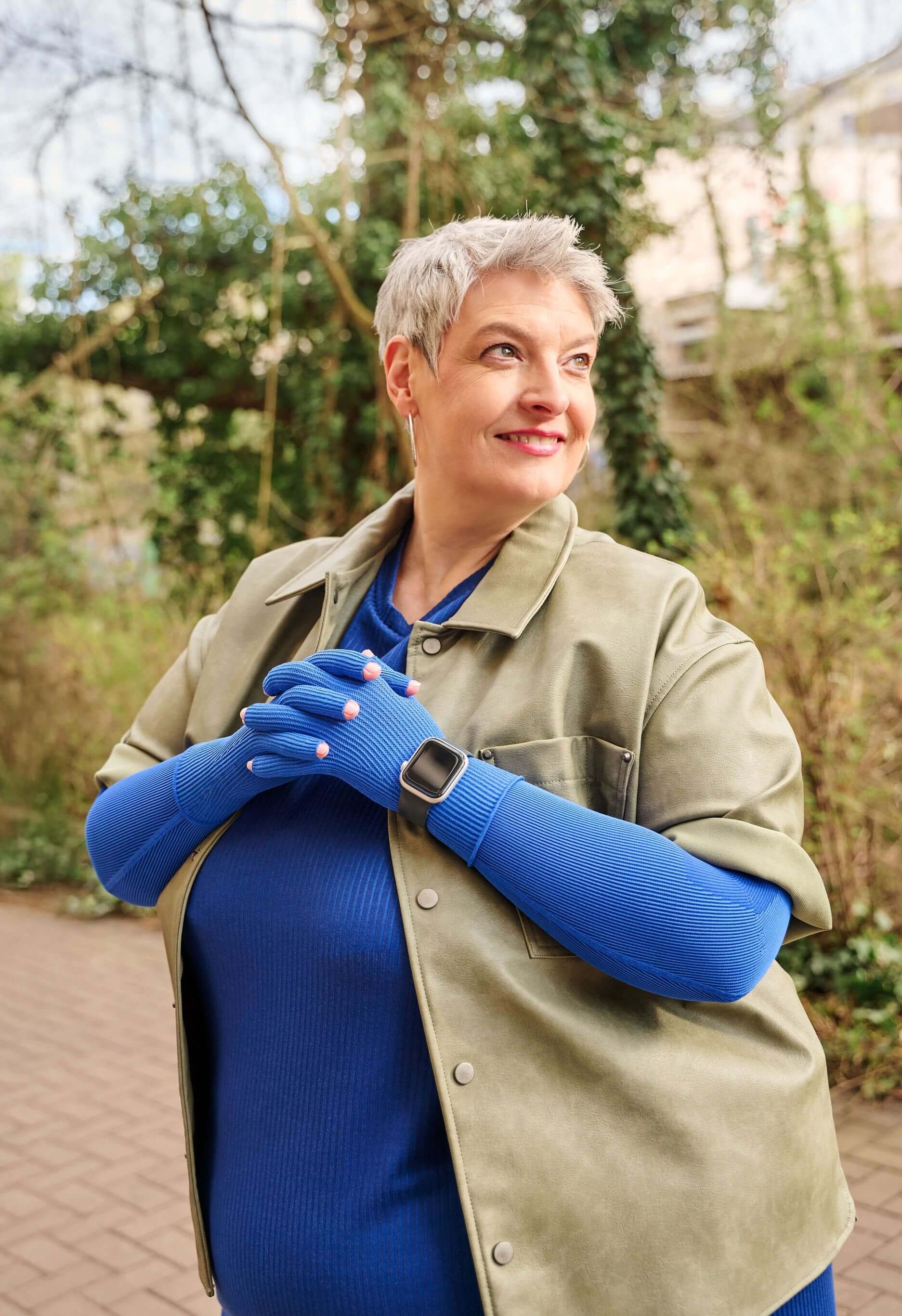Een oudere vrouw met kort grijs haar staat buiten op een geplaveid pad en lacht opzij. Ze draagt een blauw bovenstuk met blauwe handschoenen, een olijfgroen jasje en een smartwatch; op de achtergrond bomen en klimplanten.