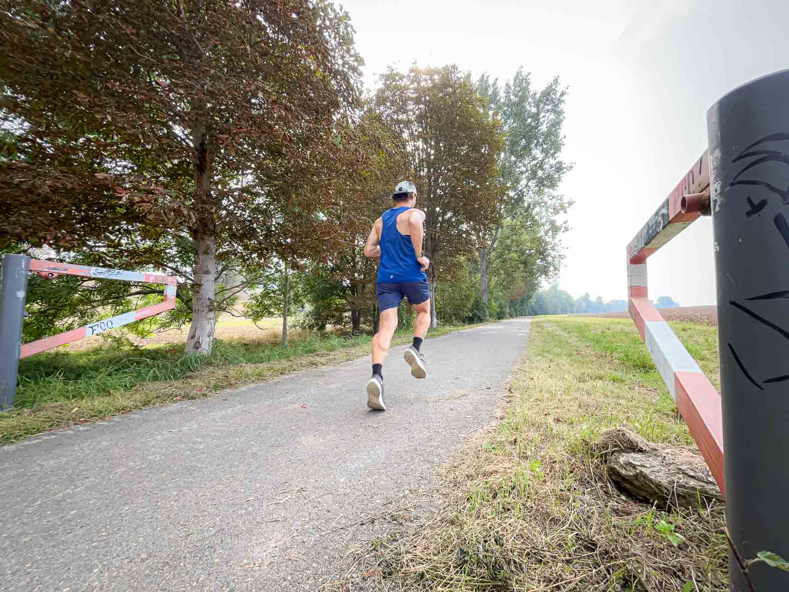 Een loper in een blauw shirt rent op een geasfalteerd pad in de natuur. Het pad wordt omgeven door bomen en een rood-witte barrière.