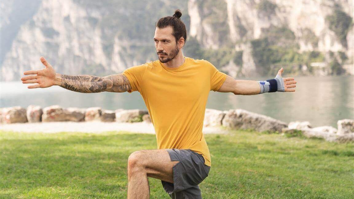 Op de foto zie je een jonge man bij een meer die een yogahouding aanneemt en daarbij een ManuTrain polsbandage draagt om pijn in de pols te verlichten.