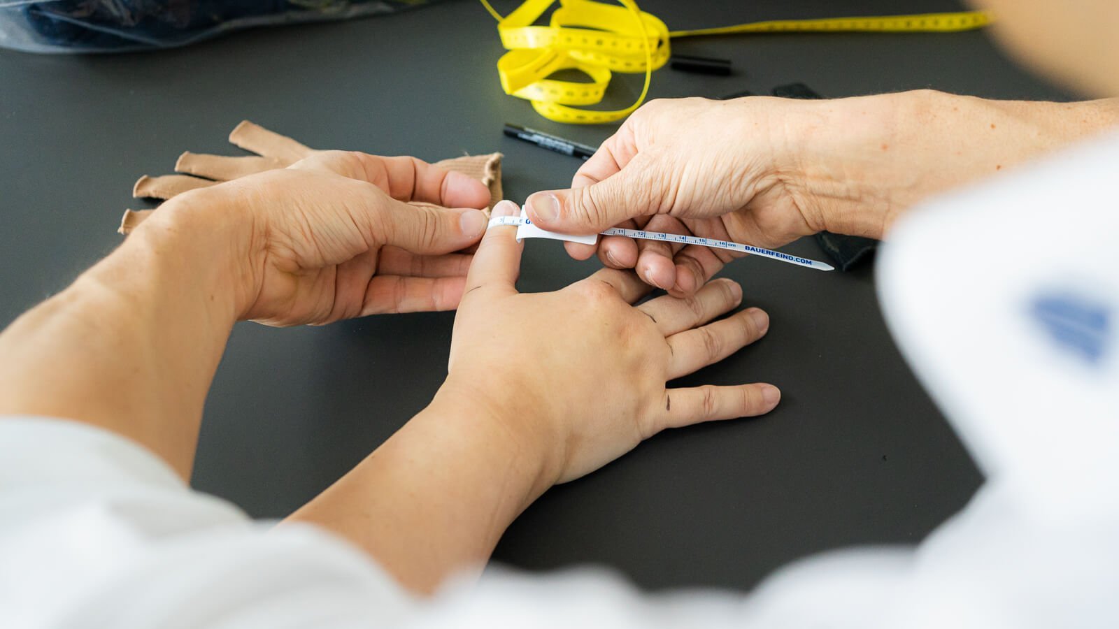 Close-up van het opmeten van Katrins hand voor een op maat gemaakte compressiehandschoen van Bauerfeind. Een persoon houdt een meetlint bij Katrins duim, terwijl haar hand plat op een donkere tafel ligt.