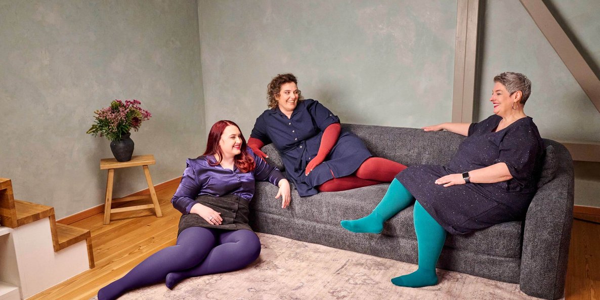 Drie vrouwen zitten ontspannen op een donkergrijze bank in een moderne woonkamer. Ze dragen kleurrijke panty's in paars, rood en turquoise bij donkere jurken of rokken en kletsen vrolijk met elkaar.