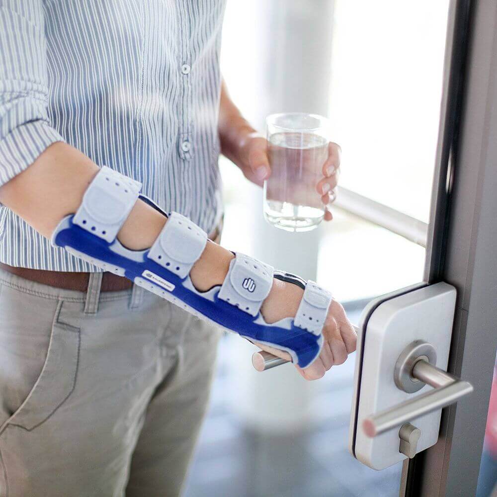 Beelduitsnede van een man die een glazen deur opent. Hij draagt aan zijn rechterarm een ManuLoc Long, een stabiliserende polsorthese, die onder andere bij een carpaletunnelsyndroom wordt gebruikt.