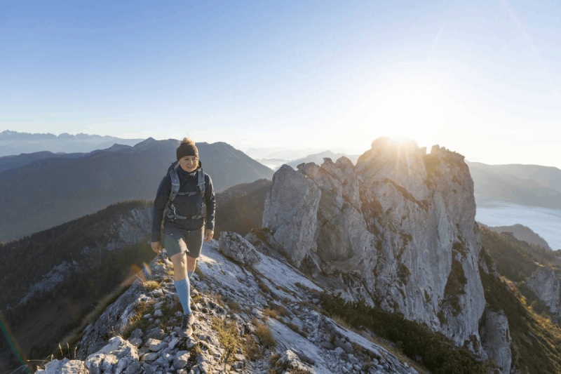 Een wandelaar met Outdoor Merino Compression Socks loopt bij zonsopgang op een smal bergpad. Deze compressiesokken bieden optimale ondersteuning en comfort voor uitdagende wandelingen.
