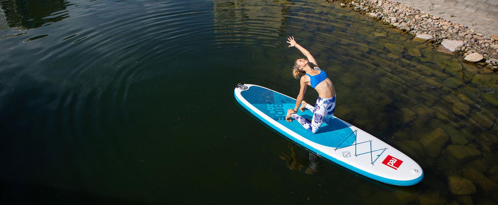Vrouw maakt yoga -oefening op het sup -bord nabij de kust onder water, een paar stenen zijn zichtbaar