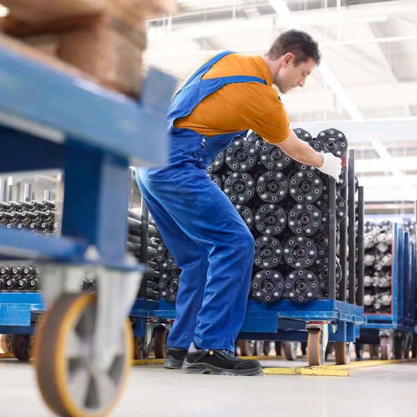Het beeld toont een jonge man in werkkleding die een steekwagen in een productiehal voortduwt. Bij lichamelijke belasting is het belangrijk om de gewrichten steeds weer te ontzien en eentonige bewegingen af te wisselen.