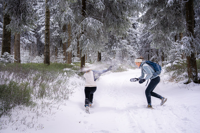 Twee vrouwen in warme winterkleding hebben een sneeuwballengevecht in een dik besneeuwd bos. De vrouw rechts draagt een grijsblauwe jas en een rugzak en gooit net een sneeuwbal.