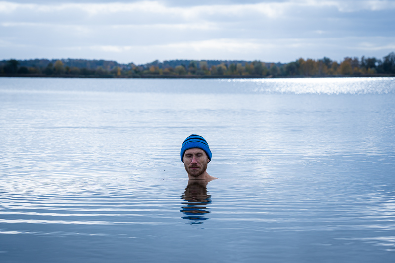 Een man staat in een rustige meer, ondergedompeld tot aan zijn nek, met een blauwe muts op, en de herfstlandschap weerspiegelt in het water.