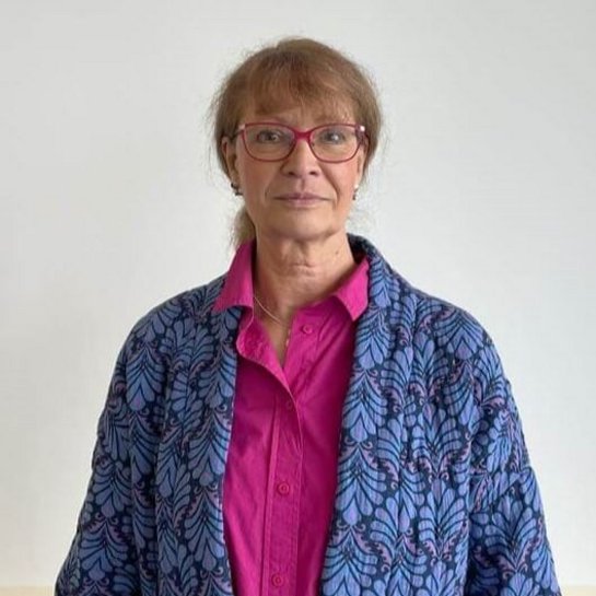 Portret van psychologe Bettina Kappe, lachend in de camera kijkend, met een rode bril, een roze blouse en een blauwe gedessineerde blazer voor een neutrale achtergrond.