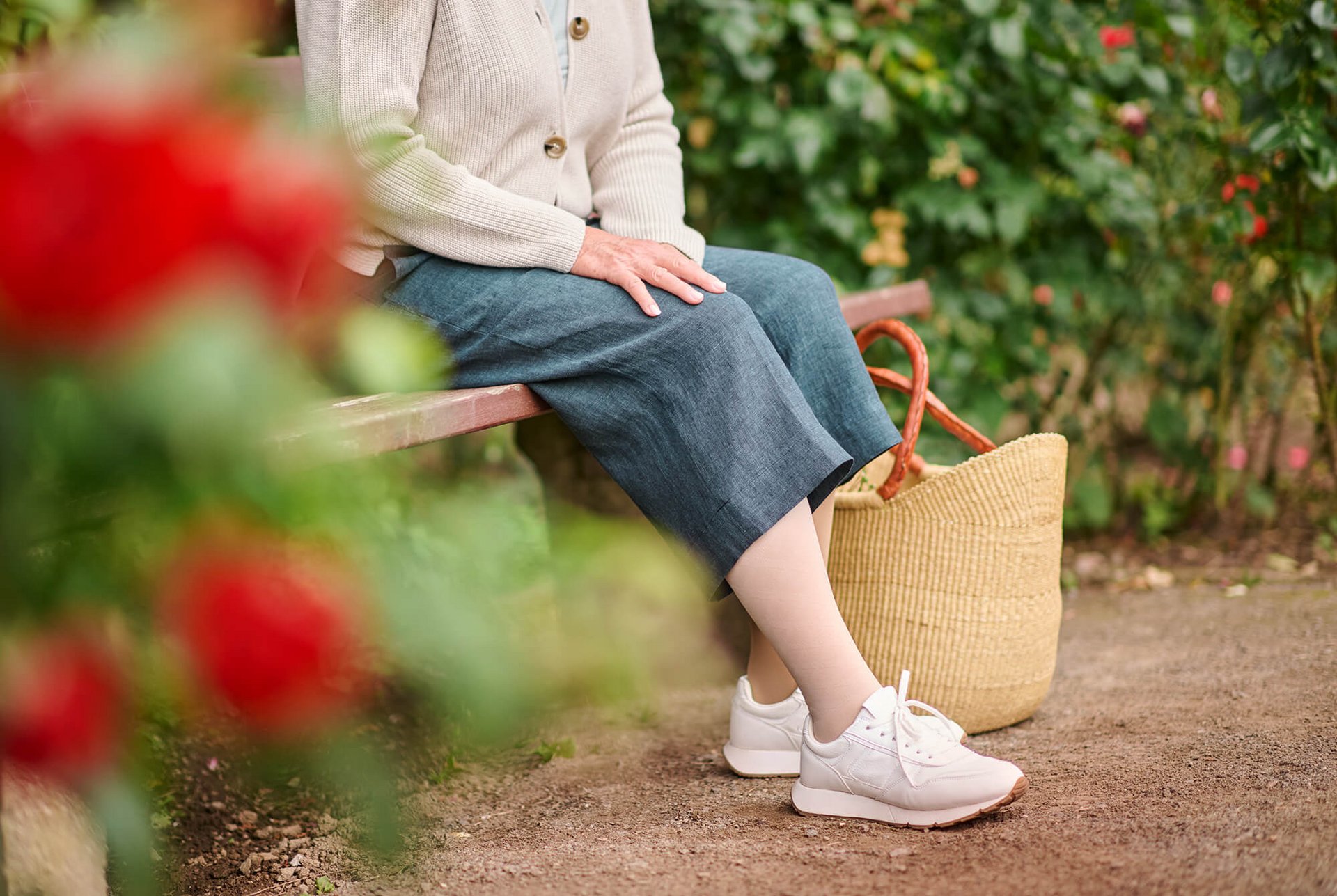 Een oudere vrouw zit ontspannen op een parkbankje. Ze draagt witte sneakers en compressiekousen uit de VenoTrain Special-lijn, omringd door bloeiende planten.
