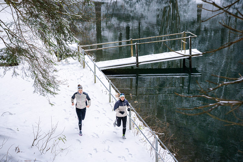 Twee hardloopsters rennen langs een besneeuwd pad naast een meer met een met sneeuw bedekte steiger. Het wateroppervlak van het meer weerspiegelt de omliggende bomen. Beide hardloopsters dragen lichte winterloopjassen en zwarte broeken.