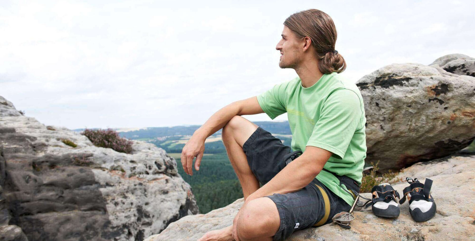 Ein junger Mann, welcher seine Haare zu einem Zopf zusammengebunden hat, sitzt auf einem Felsvorsprung und schaut in die Ferne. Er hat eine Klettersicherung um seinen Rücken gebunden. Seine Kletterschuhe stehen links neben ihm.