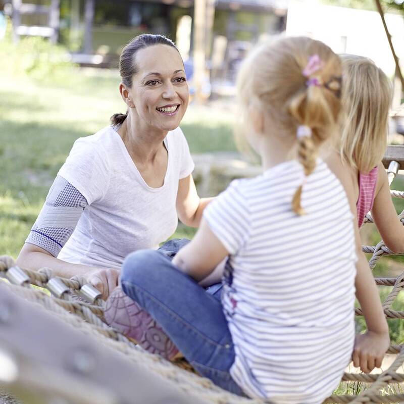 Vrouw draagt Bauerfeind OmoTrain schouderbrace tijdens spelen in het park met kinderen.