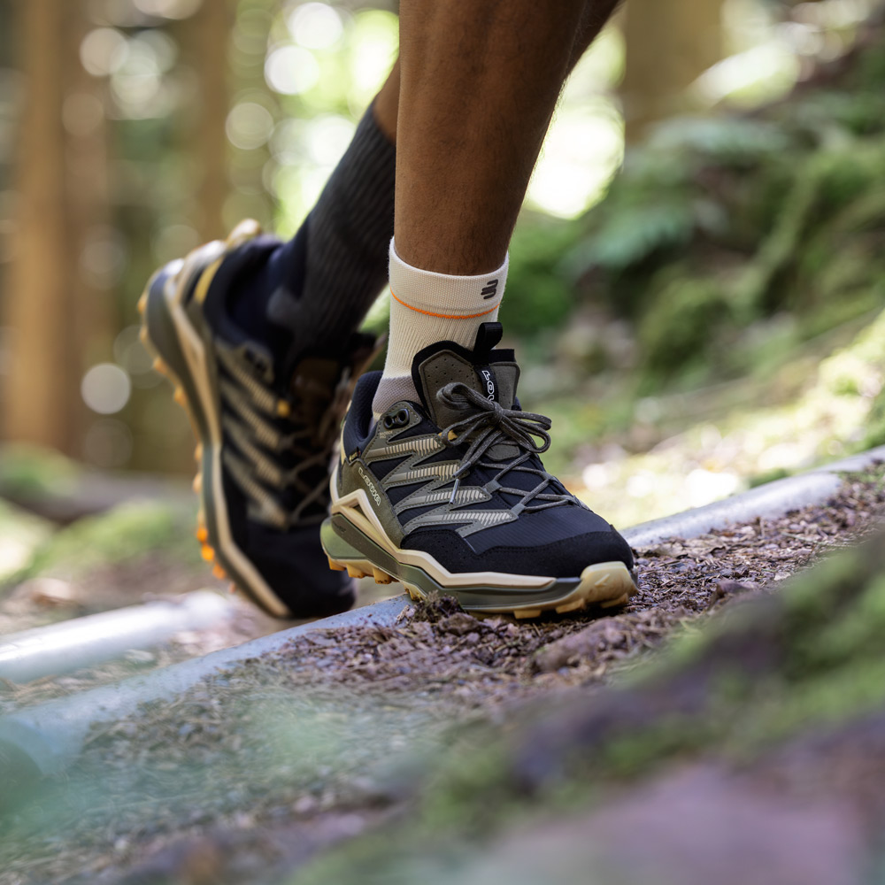 Close-up van een wandelschoen op een bosgrond met boomwortels. Boven de schoen draagt de wandelaar de Outdoor Compression Ankle Support, die gerichte compressie rond de enkel biedt en ondersteuning geeft bij elke stap.