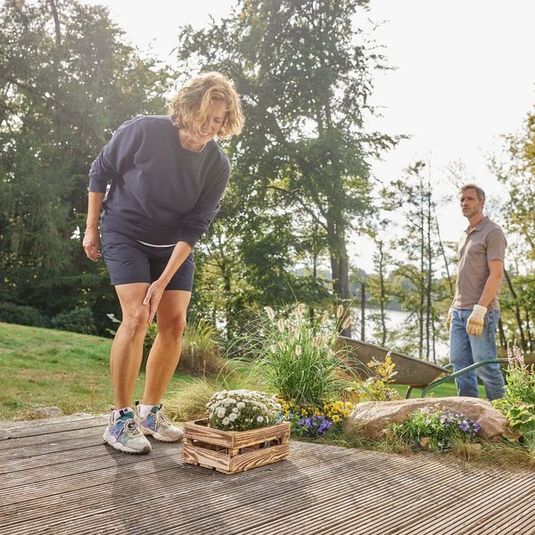 Een vrouw grijpt tijdens het tuinieren naar haar pijnlijke knie. Op de achtergrond staat een man met handschoenen toe te kijken. De tuin wordt gekenmerkt door kleurrijke bloemen en een idyllische omgeving met bomen en een meer.