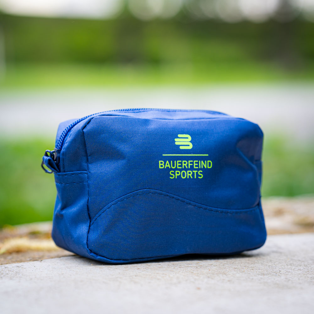 Een kleine, blauwe cosmetische tas van Bauerfeind Sports met rits ligt op een betonnen ondergrond buiten. De tas toont het Bauerfeind-logo en is compact voor onderweg.