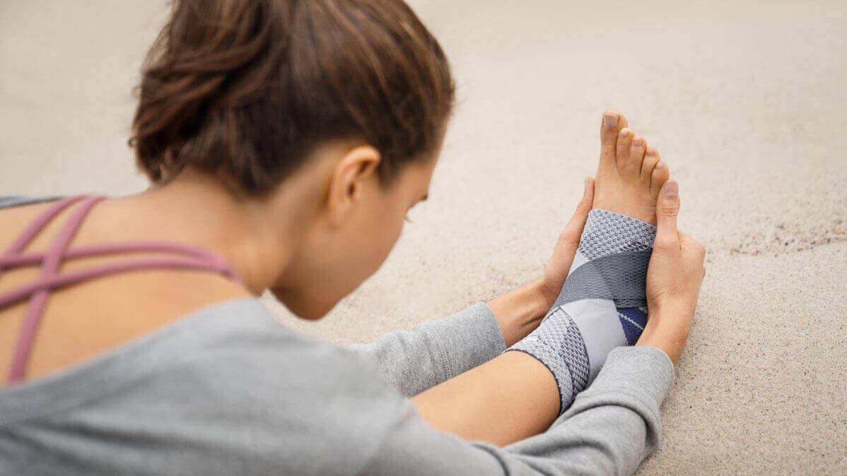 De foto toont een vrouw die op het strand in een yogahouding verblijft. Tegen pijn in de enkel draagt ze de MalleoTrain S Plus bandage van Bauerfeind.