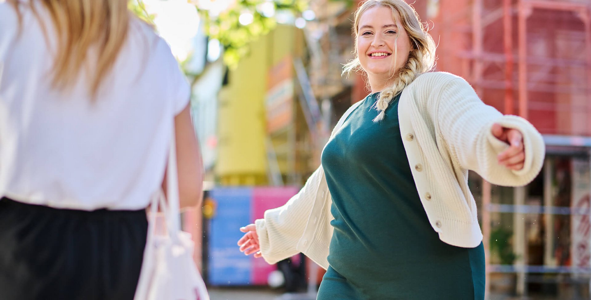 Twee vrouwen ontmoeten elkaar op een zonnige straat in een stad. Een vrouw met blonde vlecht, groene jurk en wit vest spreidt vrolijk haar armen en lacht de andere vrouw toe. Op de achtergrond zijn kleurrijke steigers en gebouwen te zien.