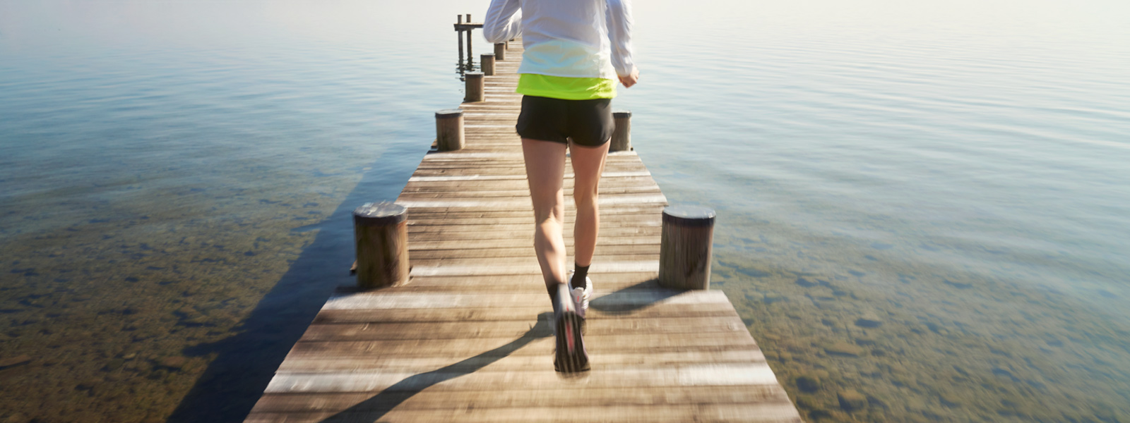 Vrouw met een wit hardloopjasje en korte zwart lopende broek loopt over een voetgangersbrug naar het water