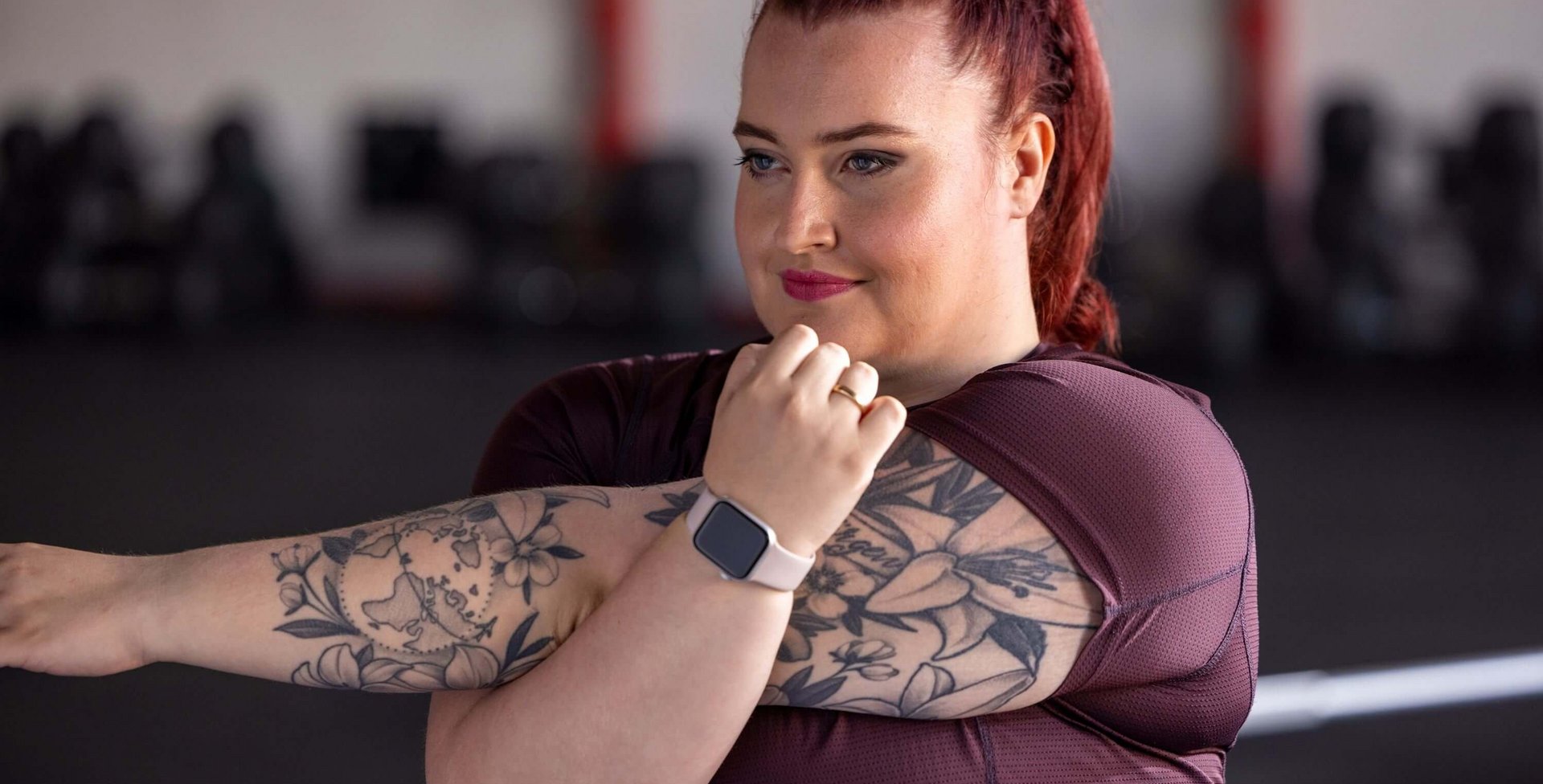 Vrouw met rood, gevlochten haar en zichtbare tatoeages op beide armen rekt haar arm in een sportschool. Ze draagt een bordeauxrood sportshirt en een smartwatch met lichtroze bandje. Op de achtergrond zijn vage fitnessapparaten te herkennen.