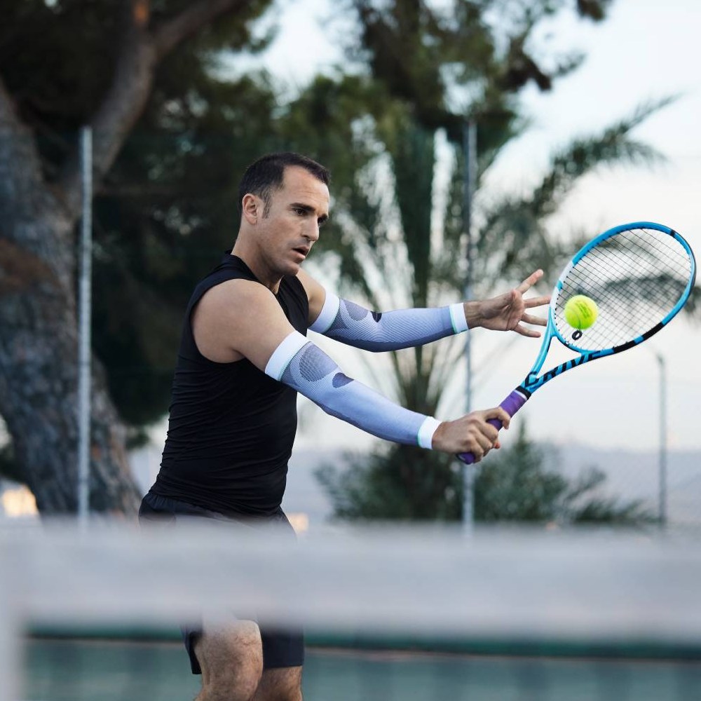 Een tennisser met witte compressiemouwen slaat een forehand. Op de achtergrond zijn bomen en een tennisbaan te zien.